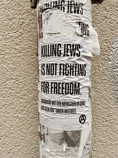 Aufkleber an Regenrinne. Weißer Hintergrund mit schwarzer Schrift in Caps:“Killing Jews is not fighting for Freedom. Solidarität mit den Menschen in Israel und den Jüd*innen weltweit.“ Darunter ein Kreis mit dem großen A.