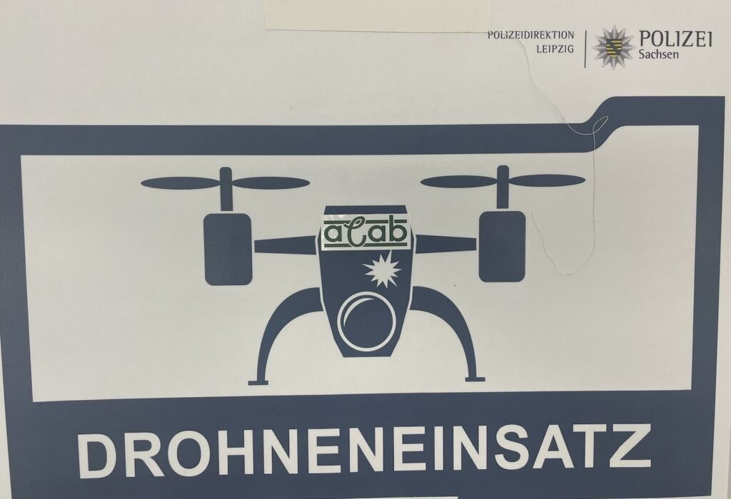 Das Bild zeigt ein blau-weißes Schild, das anscheinend von der Polizei stammt. Oben links steht "Polizeidirektion Leipzig" und rechts daneben das sächsische Polizeiwappen. In der Mitte des Schildes befindet sich eine stilisierte Darstellung einer Drohne. Auf dem Rumpf der Drohne ist der Schriftzug "ACAB" zu sehen, der von einem stilisierten Stern flankiert wird. Unter der Drohnendarstellung steht in großen blauen Buchstaben "DROHNENEINSATZ". Das Bild zeigt ein blau-weißes Schild, das anscheinend von der Polizei stammt. Oben links steht "Polizeidirektion Leipzig" und rechts daneben das sächsische Polizeiwappen. In der Mitte des Schildes befindet sich eine stilisierte Darstellung einer Drohne. Auf dem Rumpf der Drohne ist der Schriftzug "ACAB" zu sehen, der von einem stilisierten Stern flankiert wird. Unter der Drohnendarstellung steht in großen blauen Buchstaben "DROHNENEINSATZ".
